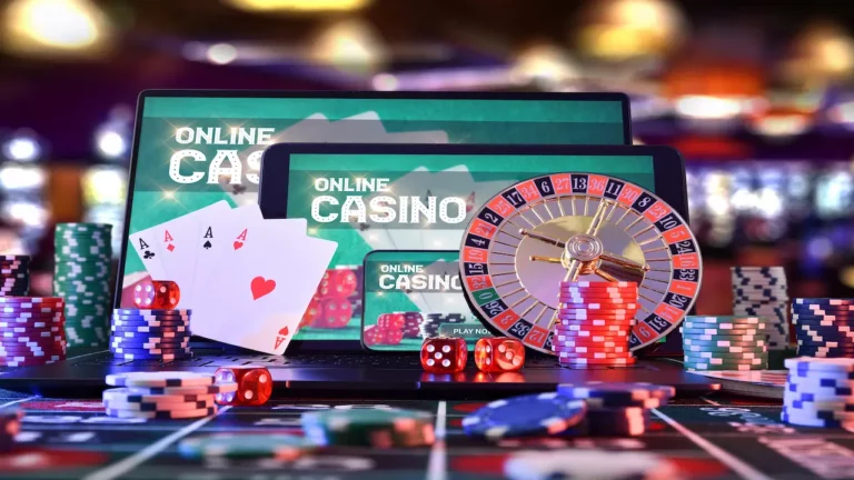 Online casino visible on electronic devices next to playing cards, roulette wheel and casino chips.